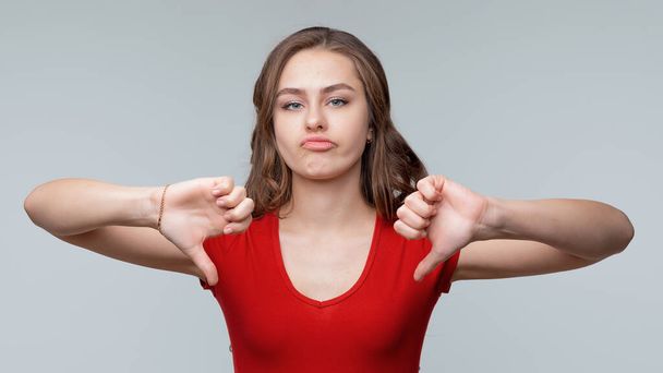 Portrait of a cute young brunette woman in red casual t shirt showing thumbs down. Gray background. Human emotions, gesture concept - Photo, Image