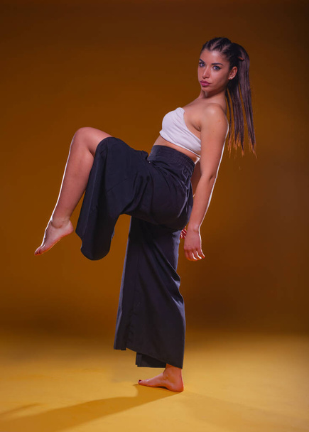 Full-body portrait of a young pretty girl posing in wide black pants and a white crop top with her leg up - Photo, image
