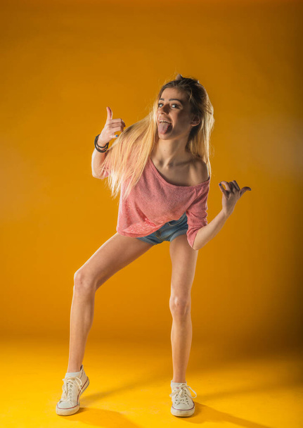 Sporty lovely girl in tank-top and denim shorts dancing with pleasure and smiling. Indoor portrait of stunning female model with vintage hairstyle waving hands on yellow background. - Photo, Image