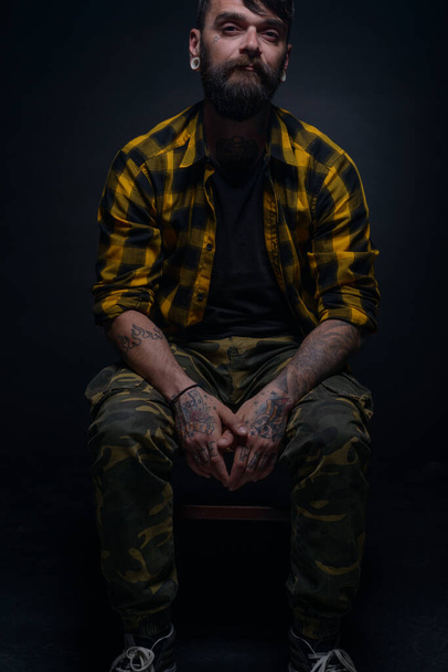 Przystojny mężczyzna model z tatuażami i koszulą tartan siedzi na krześle odizolowany na czarnym tle - Zdjęcie, obraz