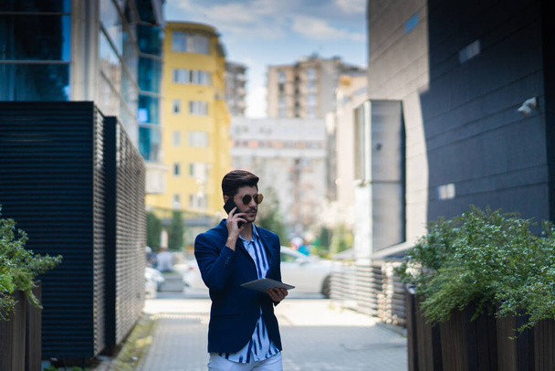 Homme d'affaires urbain parle sur son téléphone avec ses partenaires d'affaires tout en travaillant sur sa tablette et en pensant - Photo, image