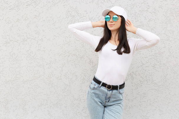 Confident pretty woman standing and looking away, expressing positive emotions, laughing happily, wearing jeans, casual shirt and baseball cap, isolated over light wall. - Photo, Image