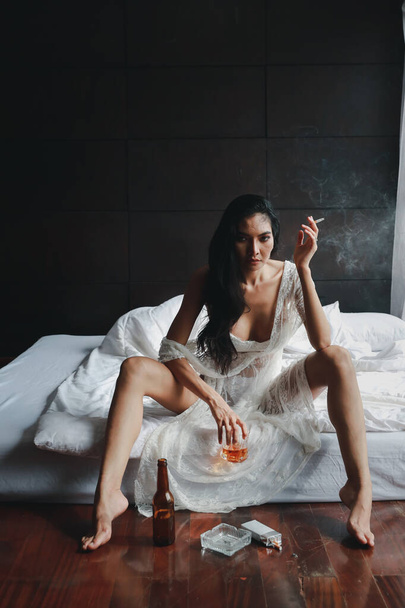 full length drunken asian woman in white lingerie, drinking and smoking while holding bottle of liquor alcohol and sitting on bed in bedroom - Foto, afbeelding