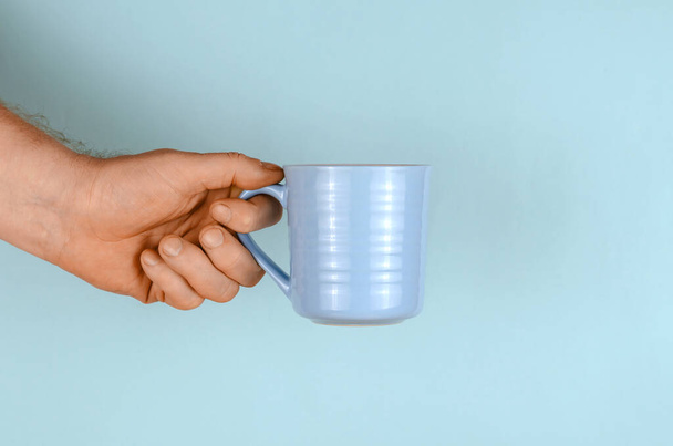 Hand with a mug on a blue background. The man is holding a light blue ceramic mug. Side view. - Foto, imagen