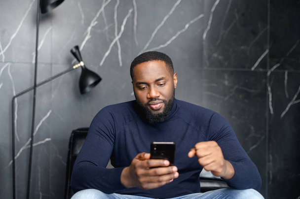 Concentrated african man sitting on couch looking at cell screen - Fotografie, Obrázek