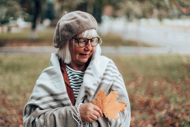 Caucasian woman in a glasses in a warm knitted blanket with a maple leaf in hands in autumn park. Cheerful lady enjoys relaxes outdoors on a warm autumn day. - Photo, Image