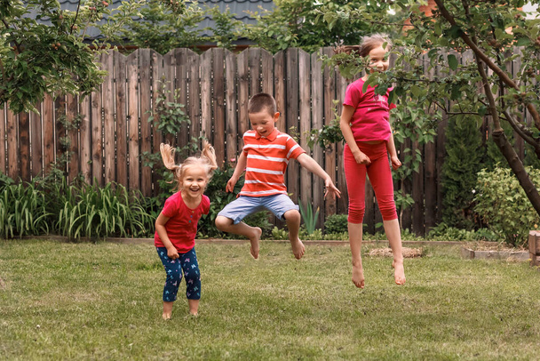 Маленькі дівчата і хлопчик сфотографувалися в стрибку на фоні саду. Троє дітей стрибають в саду на траві
.  - Фото, зображення
