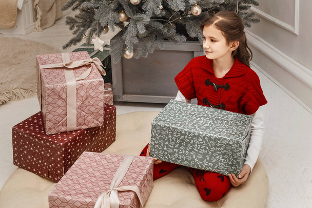 Dziewczyna w czerwonej sukience trzyma pudełka z prezentami. Dziecko siedzi obok choinki i rozpakowuje prezenty. - Zdjęcie, obraz