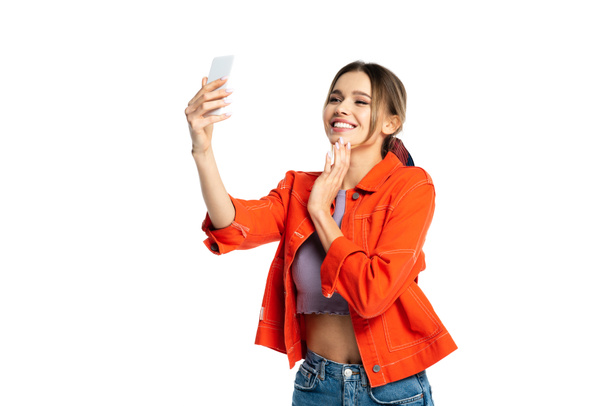 cheerful young woman in crop top and orange shirt taking selfie on smartphone isolated on white - Foto, afbeelding