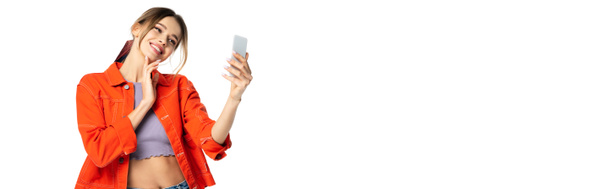 Kırpılmış bluzlu ve turuncu tişörtlü mutlu genç bir kadın beyaz ve afişte izole edilmiş akıllı telefondan selfie çekiyor. - Fotoğraf, Görsel