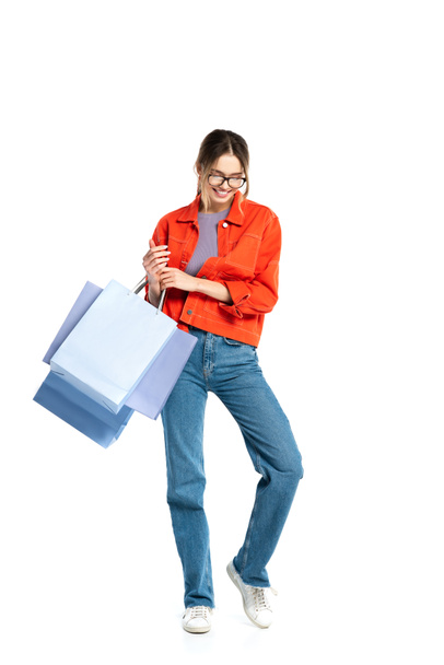 full length of pleased woman in orange shirt holding shopping bags isolated on white  - Foto, imagen