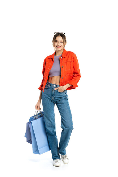 full length of happy woman in orange shirt holding shopping bags while standing with hand in pocket isolated on white  - Fotó, kép