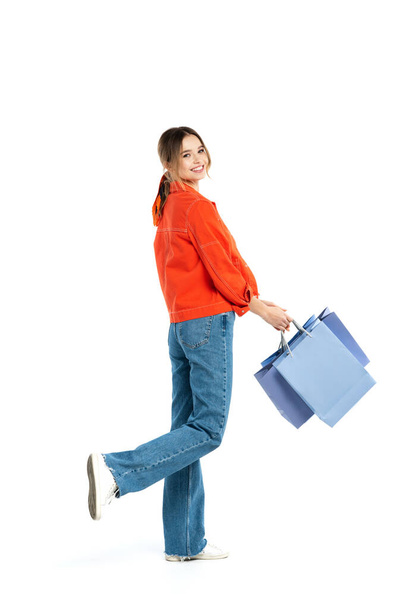 full length of pleased young woman in orange shirt holding shopping bags isolated on white  - Photo, Image