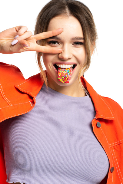 молодая женщина, торчащая языком с вкусными посыпками и показывая знак мира изолированы на белом  - Фото, изображение
