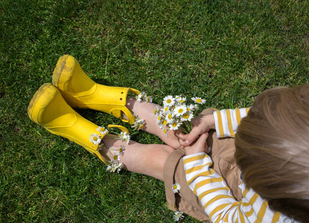 petit garçon méconnaissable dans des bottes en caoutchouc jaune vif avec un bouquet de marguerites se trouve sur l'herbe. Vue d'en haut. Un cadeau recueilli avec amour pour maman, grand-mère, copine - Photo, image