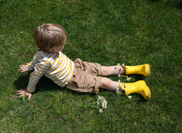 Pikkulapsi poika kirkkaan keltaisissa kumisaappaissa ja shortseissa istuu nurmikolla. Näkymä ylhäältä. Lähellä sijaitsee pieni kukkakimppu, joka on kerätty rakkaudella äidille ja isoäidille. Kesä kävelee - Valokuva, kuva