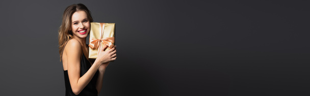 happy young woman in black slip dress holding wrapped gift box on grey, banner - Fotoğraf, Görsel