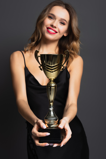 blurred happy young woman in black slip dress holding golden trophy on grey - Photo, Image