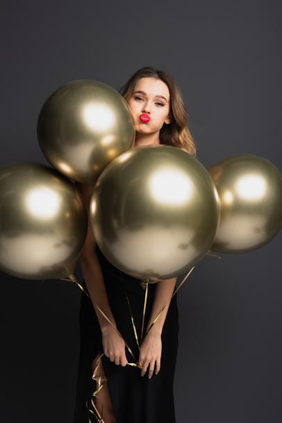 young woman in black slip dress holding golden balloons and pouting lips on grey - Fotó, kép