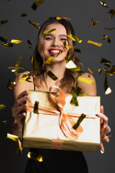 joyful young woman in black slip dress holding gift box near blurred confetti on grey - Photo, image