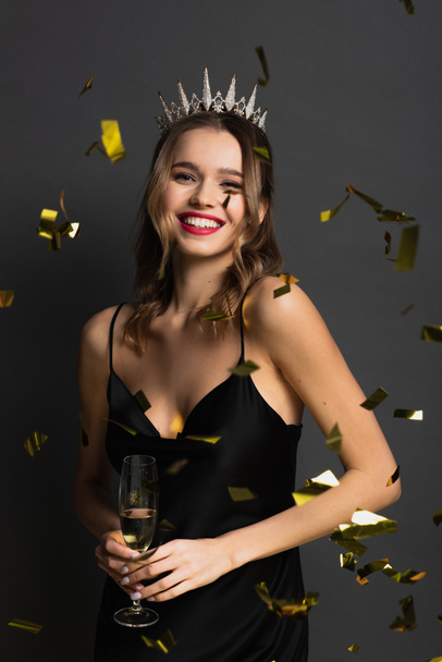 alegre joven con vestido negro y tiara sosteniendo copa de champán cerca de confeti en gris - Foto, imagen