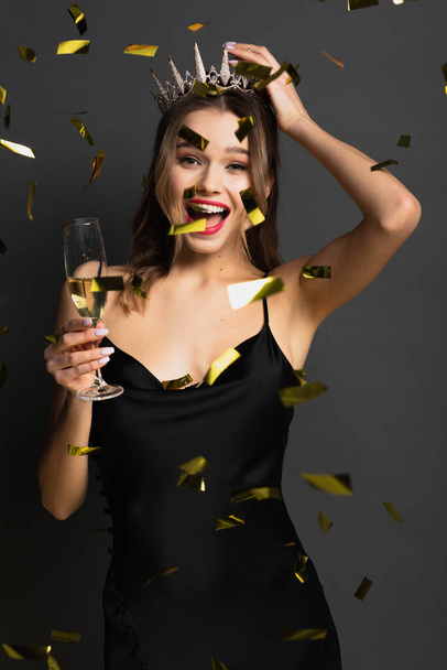 joyful young woman in black slip dress and tiara holding glass of champagne near confetti on grey - Photo, image