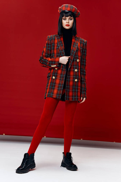 fashionable women in plaid coat and hat on red background leggings boots - Photo, Image