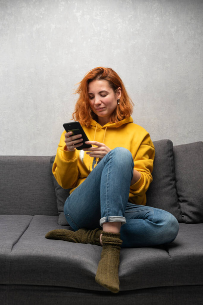 Une jeune fille sérieuse est assise sur le canapé avec un téléphone et un message de lecture. Travail à partir du smartphone. - Photo, image
