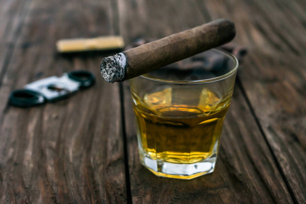  Kontrastfoto einer glimmenden Zigarre, die auf einem Glas Whiskey auf einem Holztisch liegt - Foto, Bild