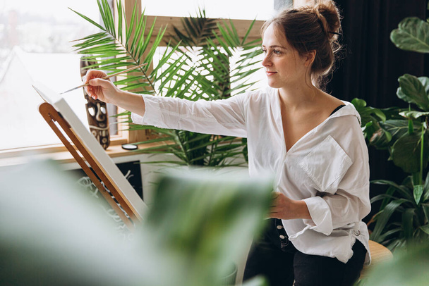 Focused young woman painting with brush on easel in studio with green plants. Inspired female artist in white shirt transferring her ideas on canvas. Creative hobby. - Foto, afbeelding