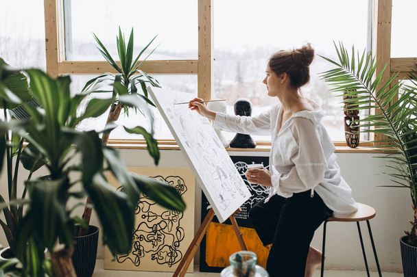 Νεαρή γυναίκα καλλιτέχνης σε λευκό πουκάμισο και μαύρο παντελόνι σχέδιο αφηρημένη εικόνα στο καβαλέτο. Ταλαντούχα γυναίκα δημιουργεί το αριστούργημά της στο στούντιο τέχνης. - Φωτογραφία, εικόνα
