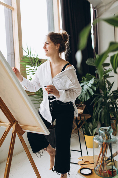 スタジオでブラシとカラーパレットでイーゼルドローイングを行う幸せな女性アーティスト。ポットの周りの緑の植物。アートセラピーとリラクゼーションの概念. - 写真・画像