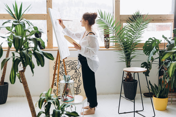 Pleasant young lady in white shirt and black pants painting with brush and color palette on easel. Talented artist creating masterpiece at bright art studio. - 写真・画像