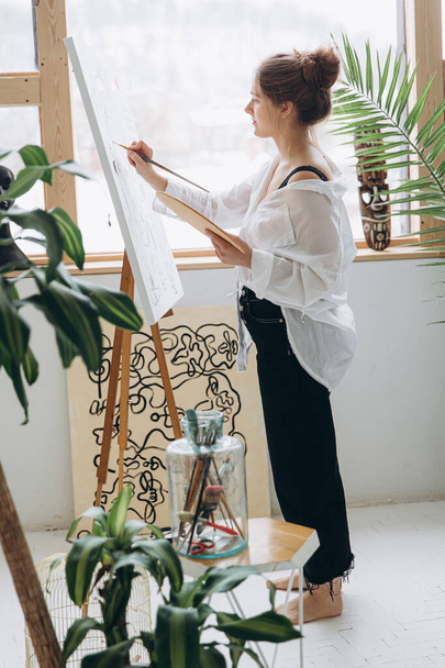 Inspirována mladá žena v neformálním oděvu, bosá stojí u velkého stojanu a maluje štětcem. Výtvarná terapie a kreativní hobby koncept. - Fotografie, Obrázek