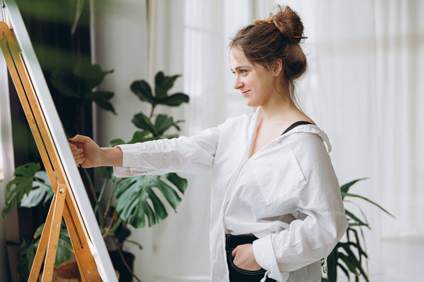 Günlük giysiler içinde, tuvale resim yapmak için fırça kullanan mutlu genç kadın. Parlak stüdyoda, saksılarda yeşil bitkilerle duran pozitif esinlenmiş sanatçı.. - Fotoğraf, Görsel