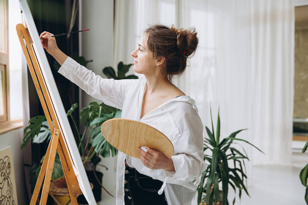Εμπνευσμένη νεαρή γυναίκα με λευκό πουκάμισο και μαύρο παντελόνι χρησιμοποιώντας πινέλο και χρωματική παλέτα. Γυναικεία ζωγραφική σε easel κατά τη διάρκεια του ελεύθερου χρόνου στο στούντιο τέχνης. - Φωτογραφία, εικόνα