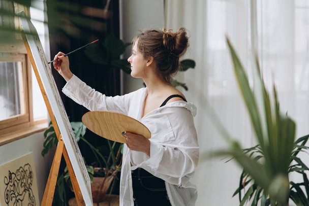 Side view of young woman in casual outfit painting on canvas with brush and color palette. Pretty artist creating art picture at bright studio. - Foto, afbeelding