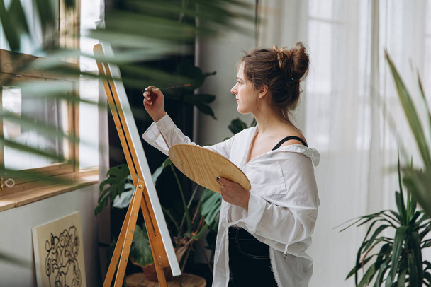 Side view of female artist in casual outfit drawing with brush on canvas. Young woman enjoying creative hobby during leisure time at art studio. - 写真・画像
