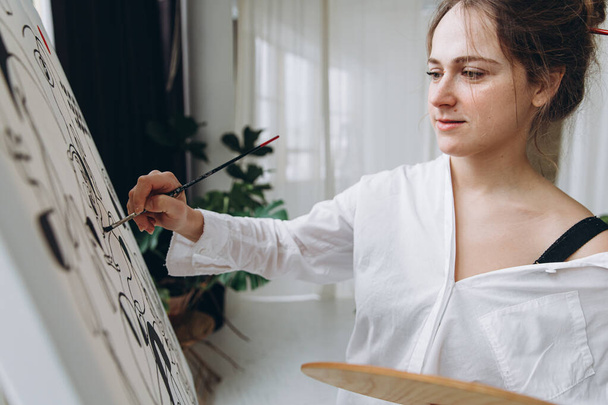 Mooie vrouwelijke kunstenaar tekening op doek met penseel en kleurenpalet. Getalenteerde vrouw in wit shirt genieten van kunsttherapie in lichte studio. - Foto, afbeelding