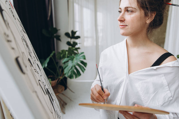 Beyaz gömlekli çekici bir kadın tuvale fırçayla resim yapıyor. Stüdyoda çizim yapan güzel bir kadın sanatçı, modern iç mimari ve yeşil bitkiler.. - Fotoğraf, Görsel