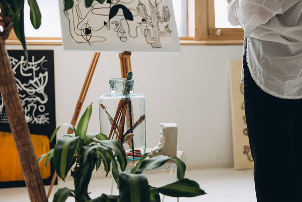 Крупним планом молода жінка малює на мольберті в художній студії. Скляна банка з різними пензлями лежить на столі. Зелені рослини на горщиках як прикраса навколо
. - Фото, зображення