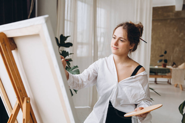 Inspired female artist in casual clothes sitting on chair in front of easel and painting with brush and color palette. Favorite creative hobby of young person. - Foto, Bild