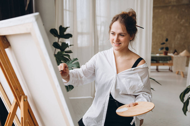 Adorable young lady in casual outfit creating drawing on easel at bright studio. Talented woman using brush and color palette for painting. - Photo, Image