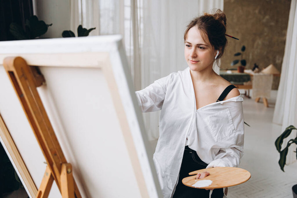 Mujer creativa joven en auriculares inalámbricos usando paleta de arte de madera mientras pinta en caballete. Artista feliz en traje casual escuchando música y dibujo sobre lienzo. - Foto, imagen