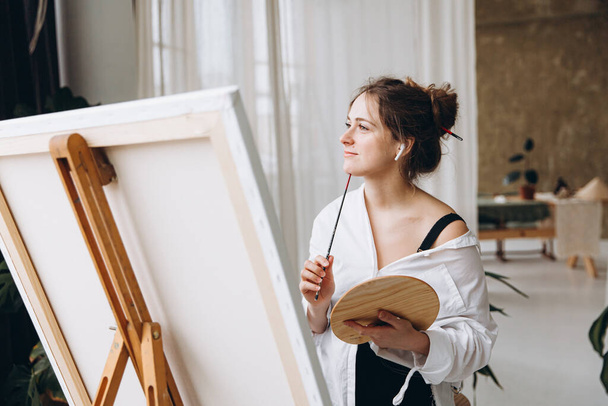 Charming woman with paintbrush and color palette in hands looking aside while searching for inspiration. Happy painter using wireless earphones for listening music during favorite hobby. - Fotó, kép