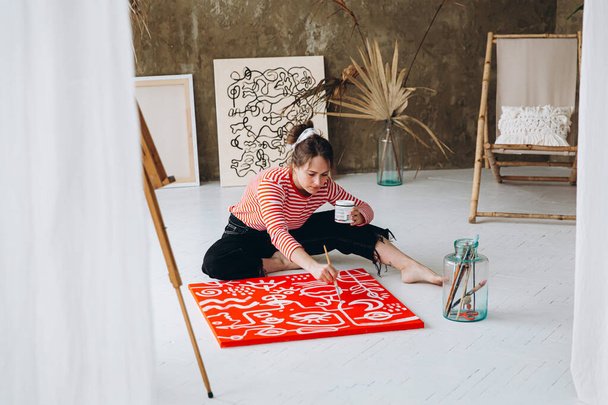 Красива молода жінка сидить на підлозі в художній студії і малює білим кольором на червоному полотні. Натхненна художниця створює абстрактний візерунок на мольберті
. - Фото, зображення