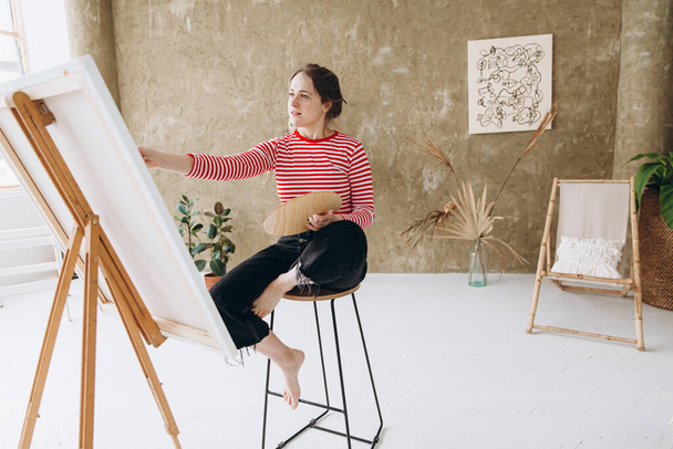 Inspired young woman painting on easel with brush and color palette at art studio. Pretty artist creating modern drawing on canvas at workplace. - Fotografie, Obrázek