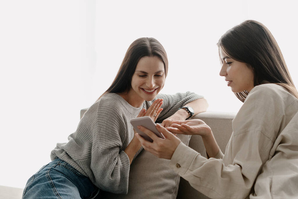 Kaksi nuorta aikuista naista, jotka katsovat älypuhelimen näyttöä, puhuvat toisilleen ja hymyilevät leveästi. Parhaat ystävät viettävät viikonlopun yhdessä, istuvat sohvalla kotona. Sosiaalinen media ja online app käsite - Valokuva, kuva