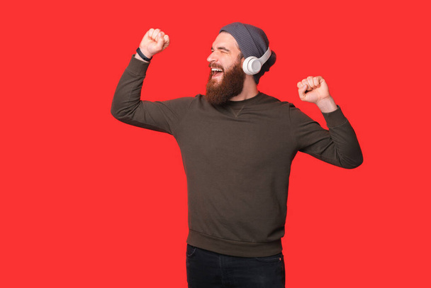 Happy cheerful bearded hipster man listening music at earphones and dancing over red background. - Foto, Imagem