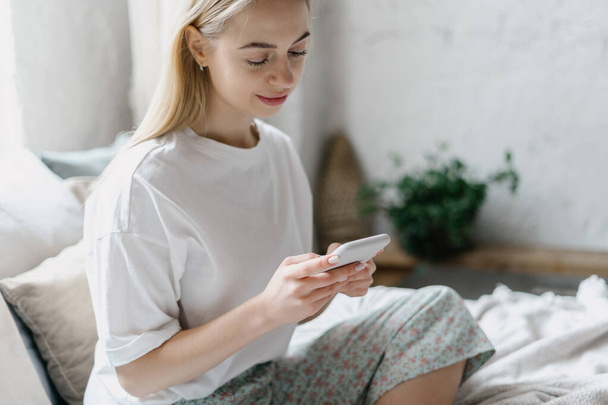 Mulher jovem calma e bonita usando o aplicativo on-line em seu smartphone moderno. Sorrindo surfe feminino na internet, sentado na cama de conforto em pijama, passando a manhã em casa em quarto acolhedor luz - Foto, Imagem